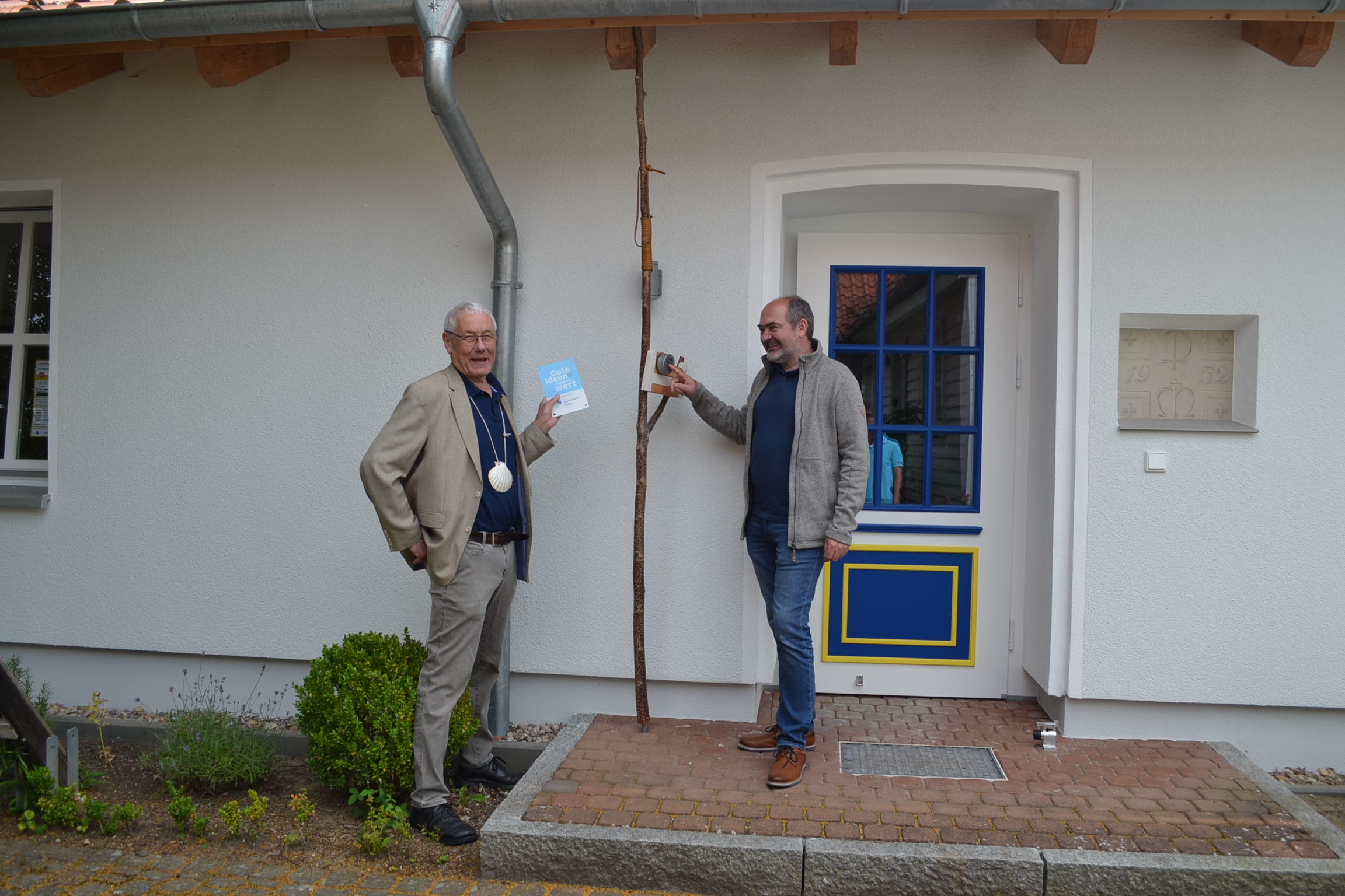 2 männliche Personen stehen vor einer Haustür. Die eine Person drückt auf die Schließanlage. Die andere Person schaut dabei zu mit einem Schild in der Hand.