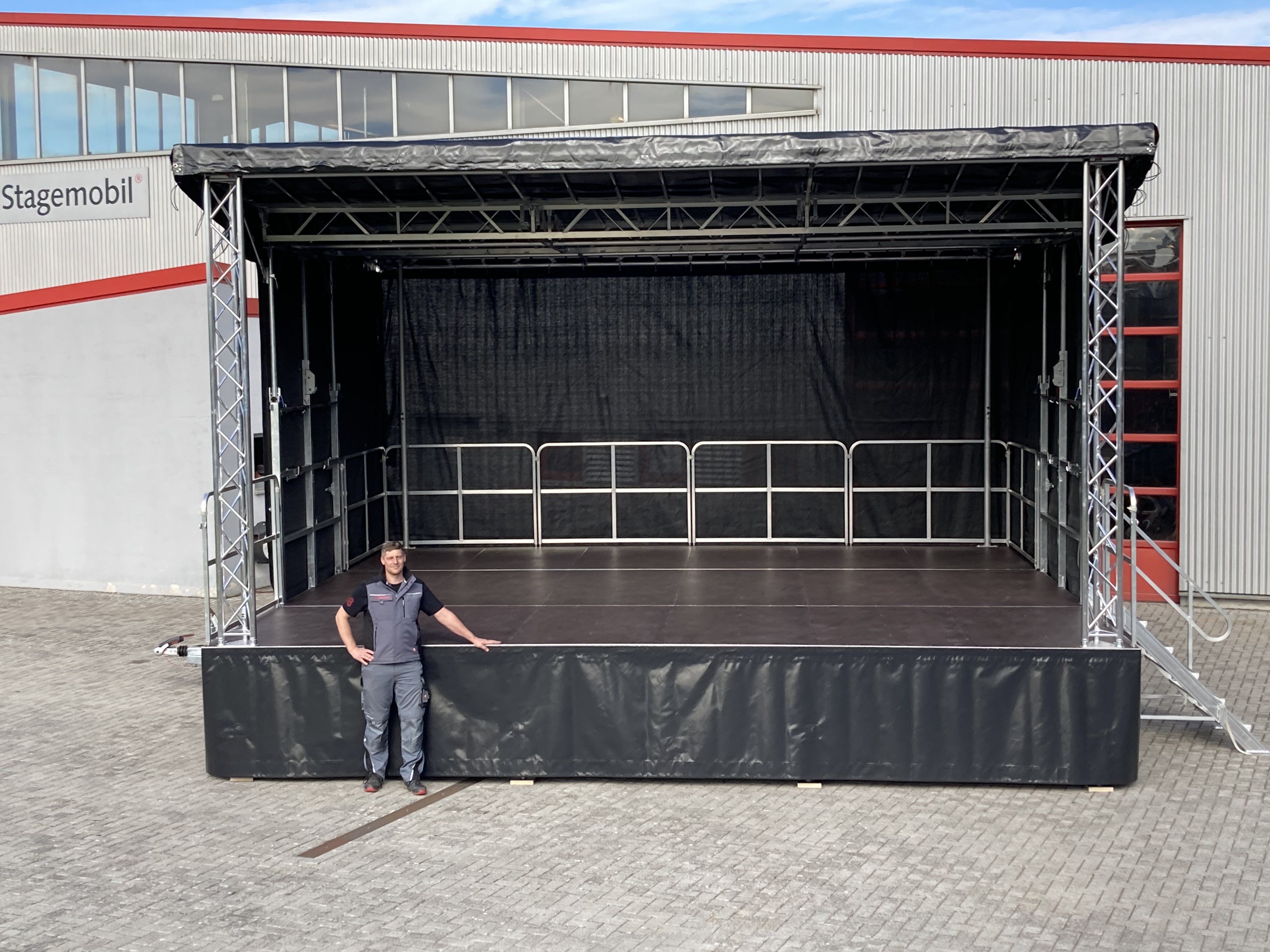 Im Vordergrund befindet sich ein mobiler Bühnenanhänger. Davor steht eine männliche Person, dessen linke Hand sich auf dem Bühnenpodest befindet. 