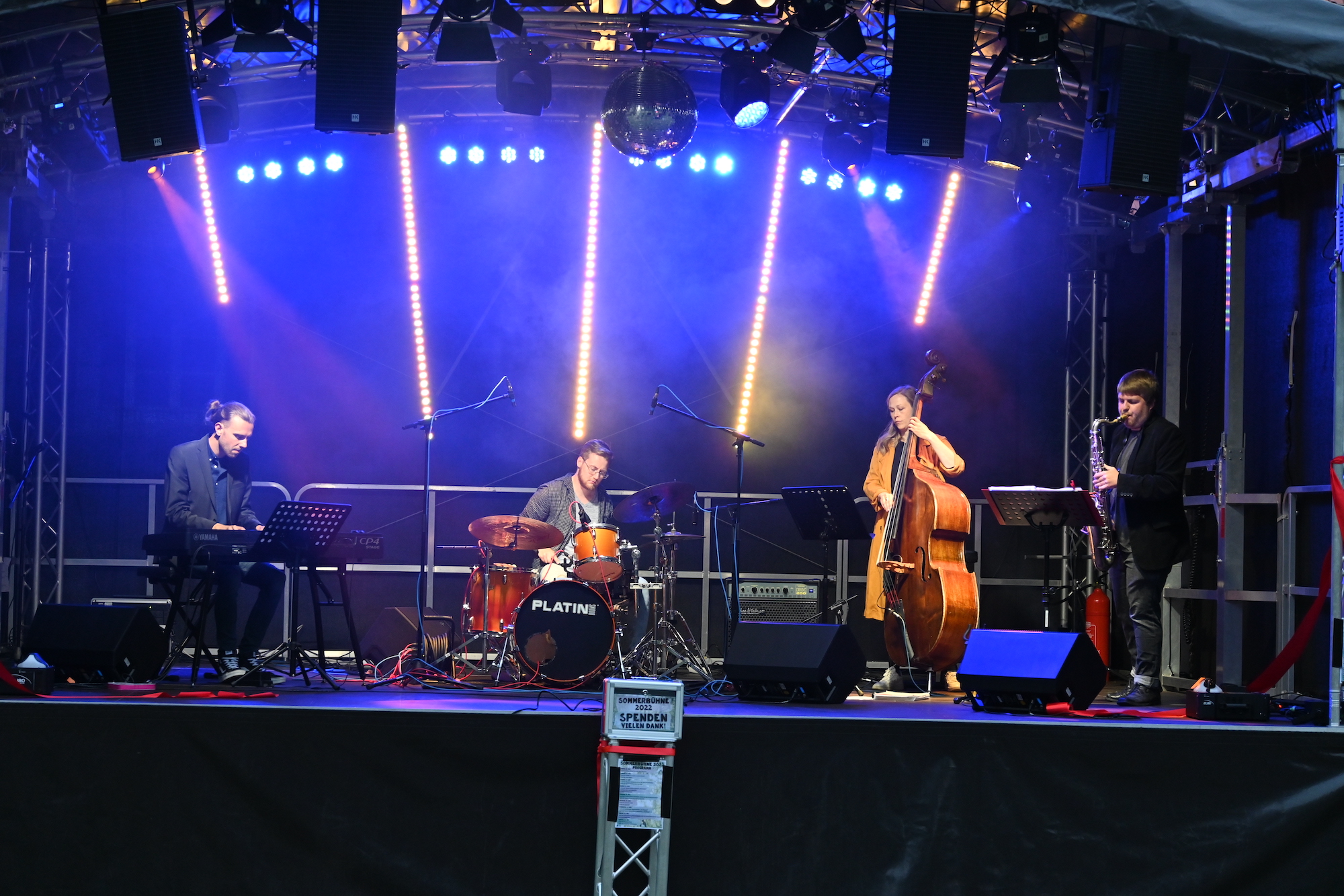 Vier Personen stehen auf einer beleuchteten Bühne und spielen jeweils auf einem Musikinstrument.