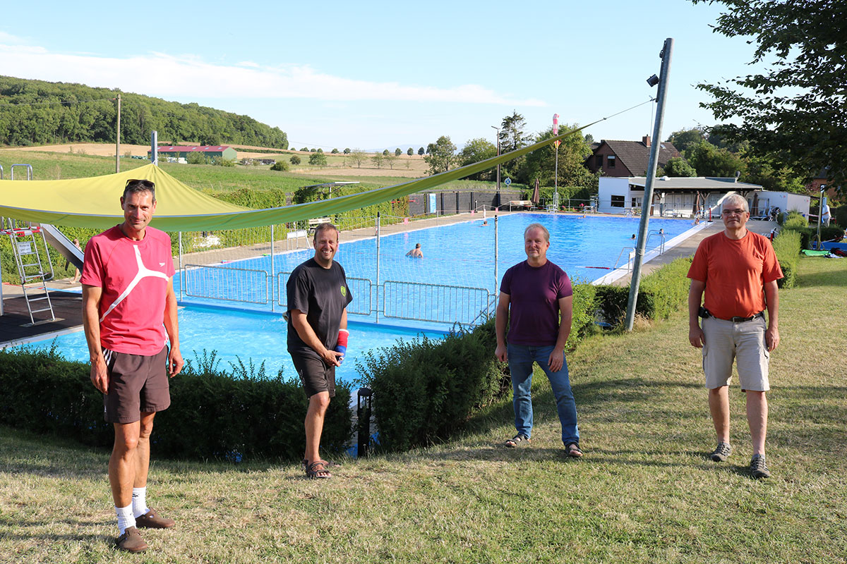 Blick auf ein großes Schwimmbecken, im Hintergrund eine grüne Landschaft im Sommer, im Vordergrund vier Personen, die in die Kamera blicken.